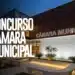 Prédio da Câmara Municipal com fachada em madeira ripada, letreiro luminoso e jardim frontal iluminado à noite.