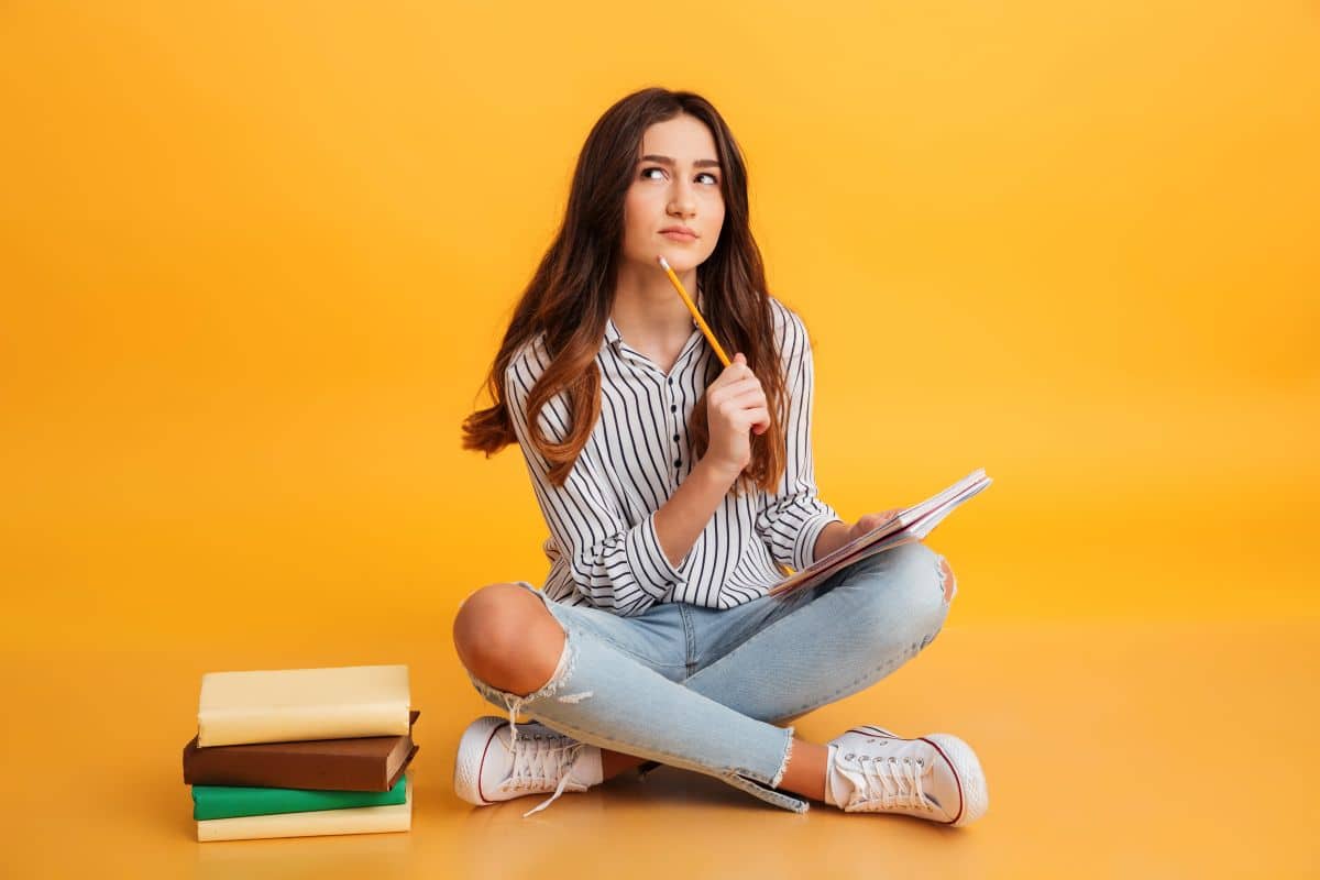 Estudante pensativa com caderno e lápis em fundo laranja.