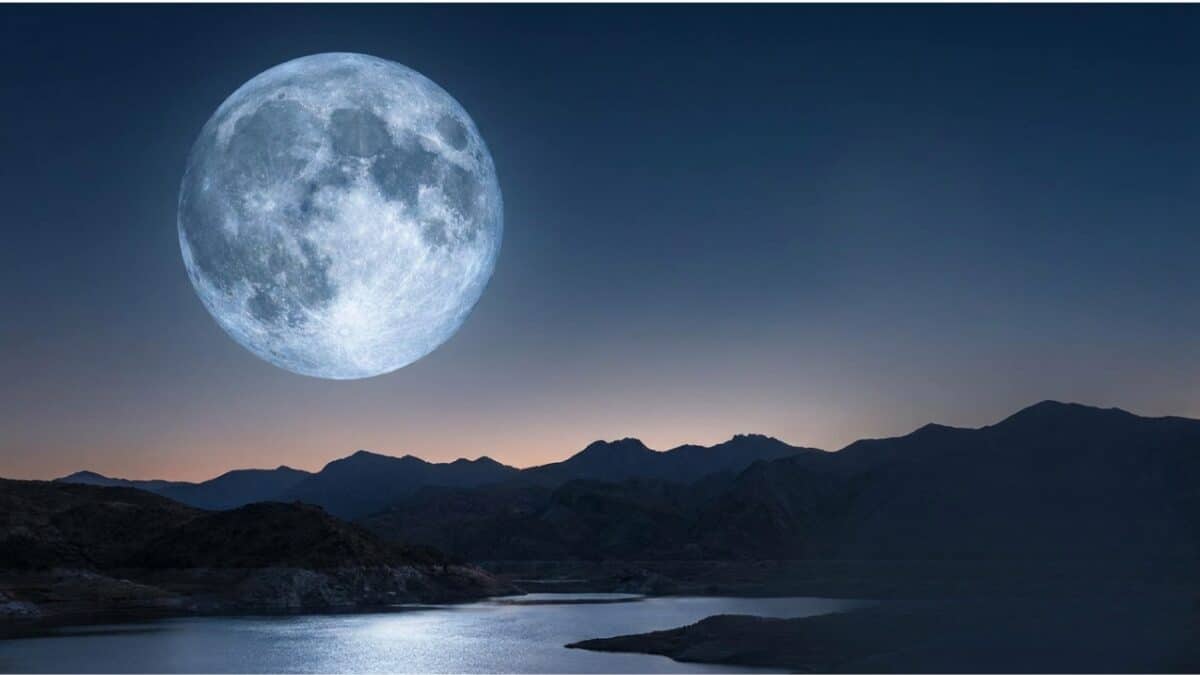 Lua cheia imensa brilha sobre lago tranquilo com reflexo prateado, cercado por montanhas escuras ao entardecer.