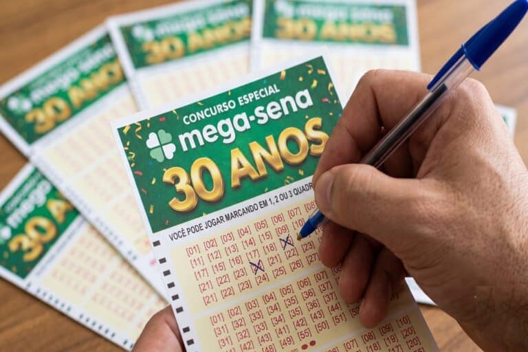 Mão segurando caneta azul marca números no volante verde do concurso especial Mega-Sena 30 Anos