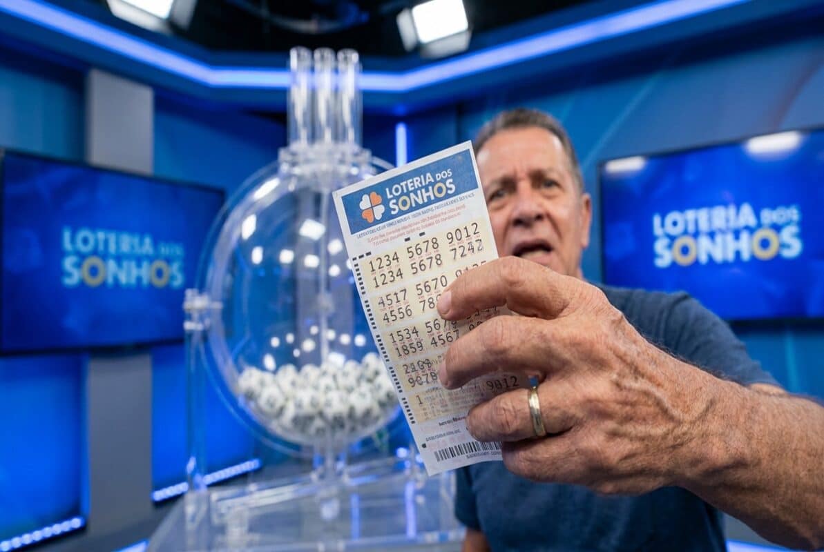 Homem de meia-idade segura dois bilhetes da Loteria dos Sonhos diante de globo com bolas brancas em estúdio azul