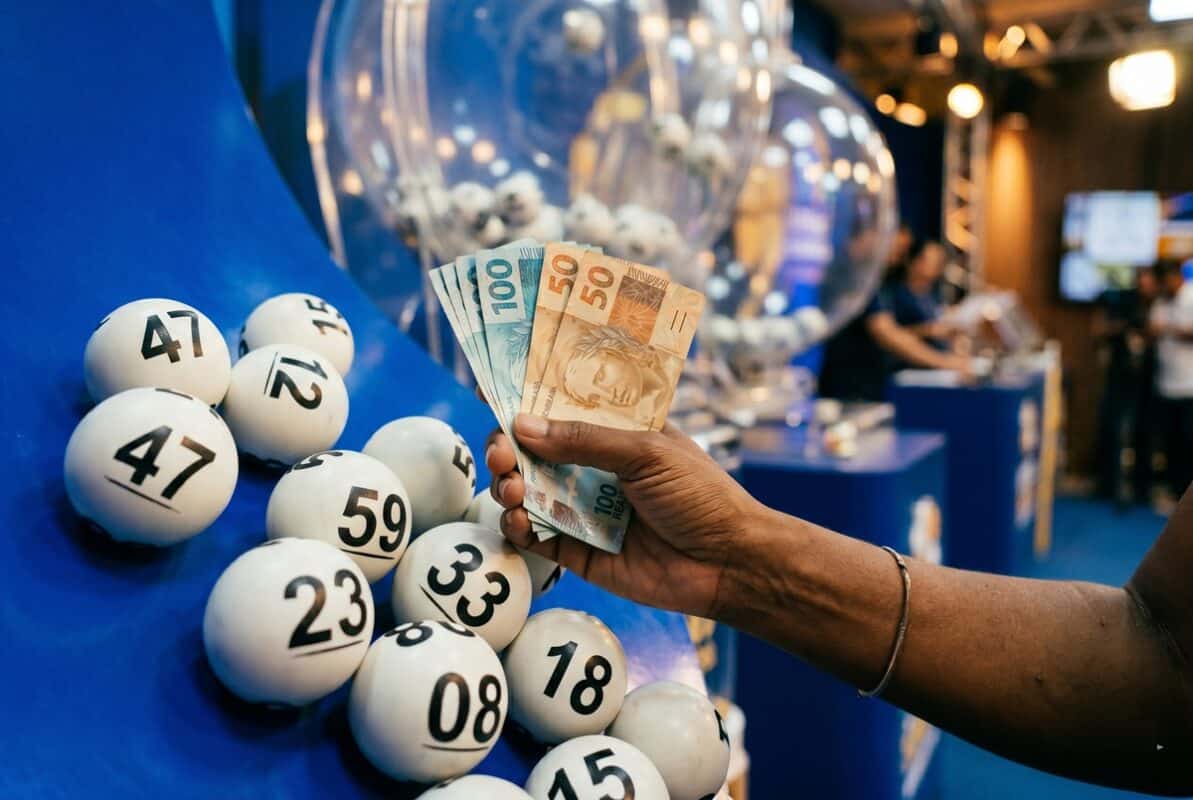 Mão segurando notas de 50 e 100 reais ao lado de bolas brancas numeradas em globo de sorteio azul