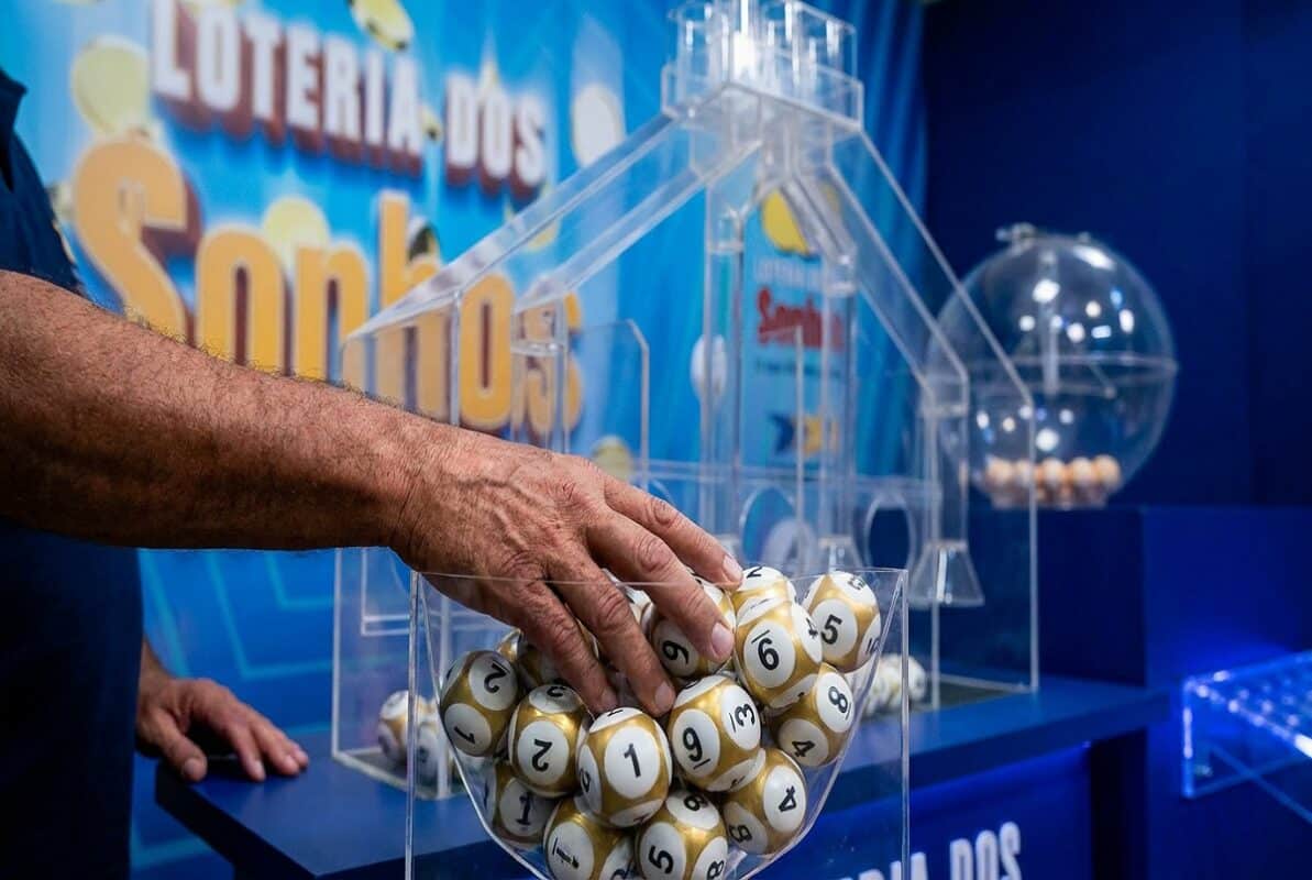 Mão masculina pegando bolas de sorteio douradas e brancas numeradas em globo de acrílico da Loteria dos Sonhos