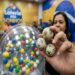 Mulher segura bolas com bichos e números sorteados da Loteria dos Sonhos, ao lado do globo colorido da LOTECE
