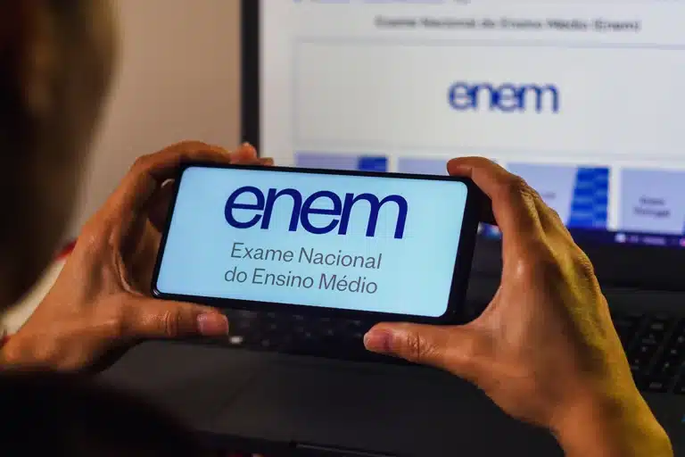 Mãos segurando smartphone com logo do ENEM na tela, em frente a monitor de computador exibindo a Página do Participante.