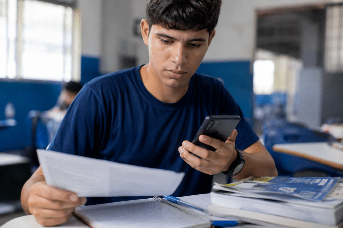 Jovem de camiseta azul-marinho sentado em sala de aula segura folha de papel na mão direita e smartphone na esquerda.