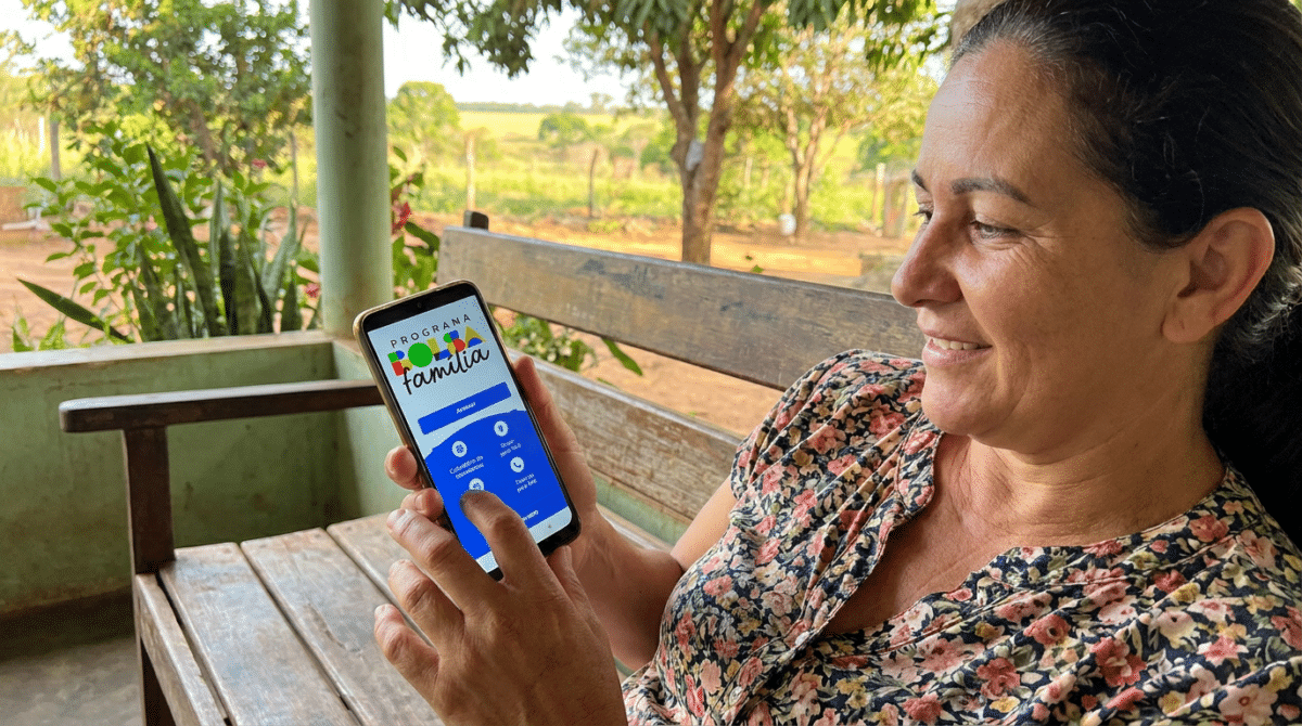 Mulher sorri ao acessar o aplicativo do Bolsa Família no celular, sentada em banco de madeira ao ar livre.