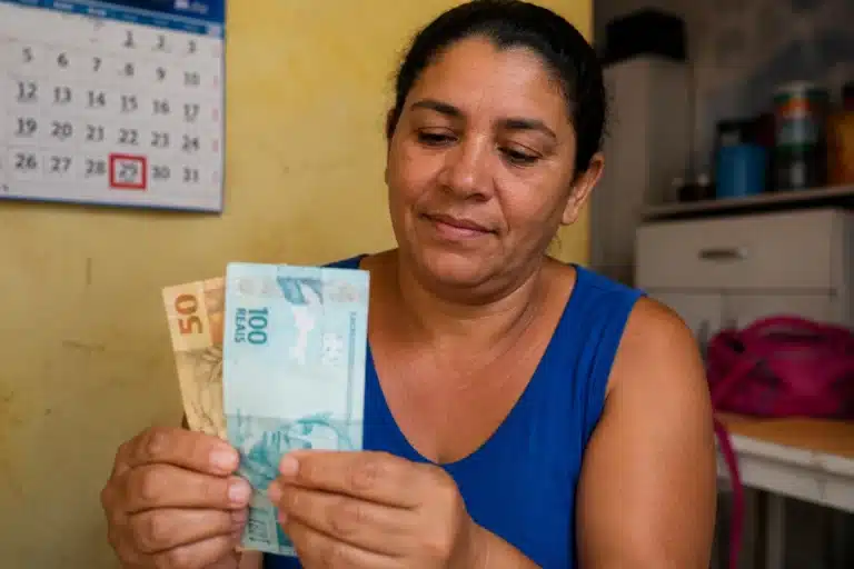 Mulher segurando cédulas de reais em ambiente doméstico, representando o pagamento final de maio do Bolsa Família.