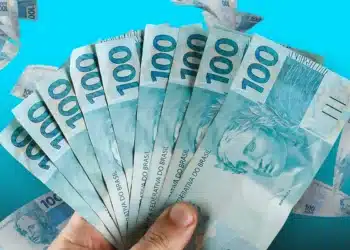 Mão segura várias cédulas de cem reais, em referência ao pagamento do Pé-de-Meia de maio de 2026