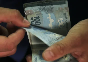 Mãos de pessoa contando cédulas de cem reais sobre fundo escuro