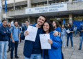 Dois jovens sorriem e exibem documentos oficiais em frente a um centro de formação enquanto outros aprovados comemoram ao fundo