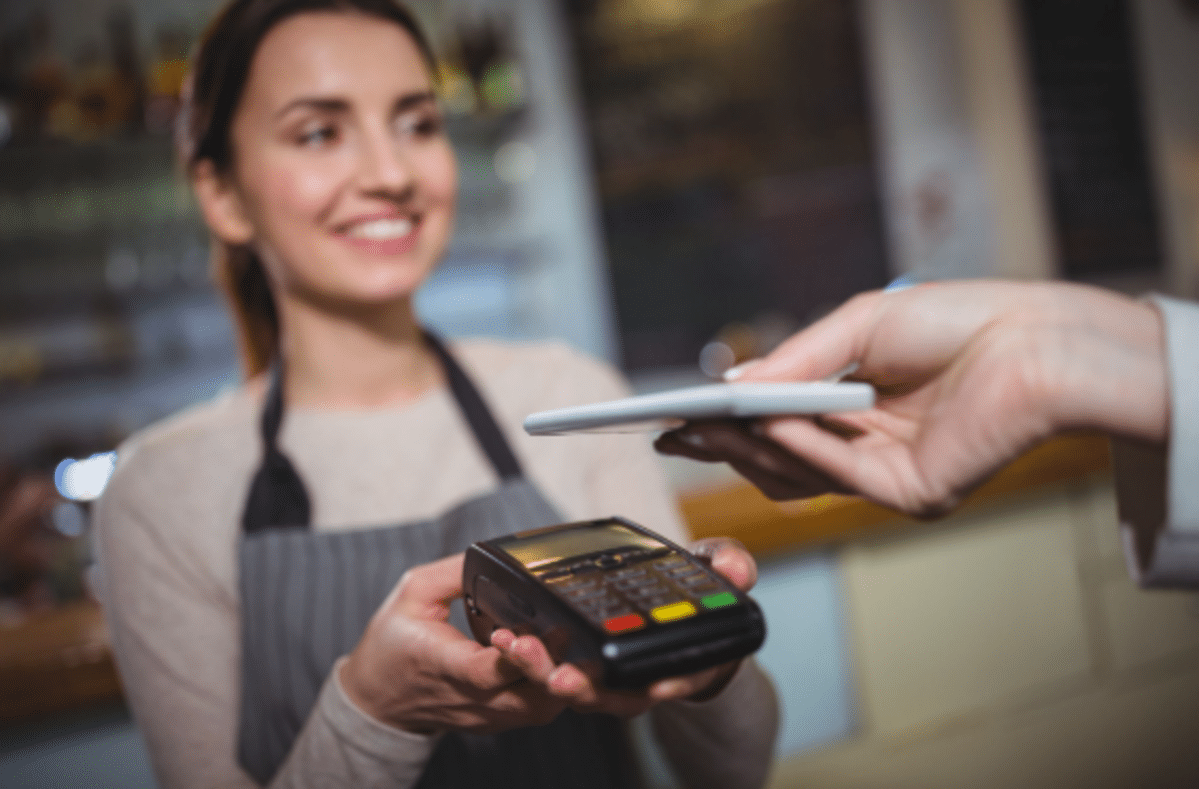 Atendente sorridente segura maquininha enquanto cliente aproxima celular para pagamento NFC