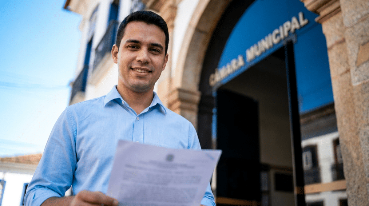 Homem jovem de camisa azul clara segura documento com brasão, sorrindo em frente à Câmara Municipal.
