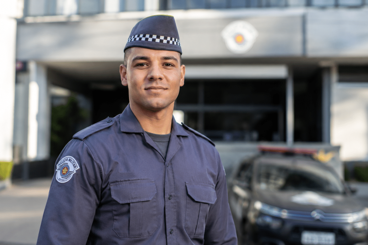 Policial militar com uniforme cinza da PMESP, quepe xadrez e brasão bordado, em frente a viatura e prédio oficial.