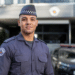 Policial militar com uniforme cinza da PMESP, quepe xadrez e brasão bordado, em frente a viatura e prédio oficial.