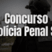 Policial penal de costas com capacete tático e uniforme preto com inscrição "PENAL", bandeiras ao fundo.