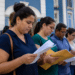 Grupo de pessoas lendo folhas e envelopes amarelos em fila externa junto a prédio colonial azul e branco.