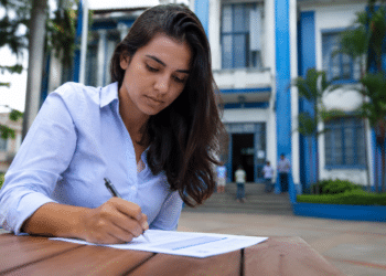 Mulher de camisa azul clara escreve em documento sobre mesa de madeira, com prédio azul institucional ao fundo.