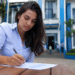 Mulher de camisa azul clara escreve em documento sobre mesa de madeira, com prédio azul institucional ao fundo.