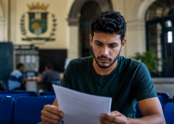 Homem de cabelos cacheados e camiseta verde escura sentado em cadeira azul lendo papel em recepção com brasão ao fundo.