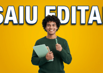 Jovem de cabelos cacheados com suéter verde segurando cadernos e caneta sorri diante de fundo amarelo com texto "Saiu Edital"