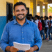 Professor de camisa azul segura folhas de papel enquanto sorri, com alunos uniformizados ao fundo em pátio escolar.