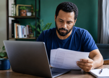 Homem de barba com camiseta azul marinho analisa papéis em frente a notebook cinza em escritório doméstico.