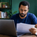 Homem de barba com camiseta azul marinho analisa papéis em frente a notebook cinza em escritório doméstico.