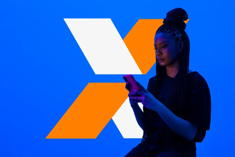 Mulher usa celular diante de marca em formato de X laranja e branco que representa o programa Desenrola.