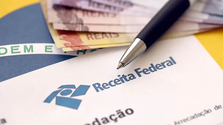 Folha com timbre da Receita Federal repousa sobre superfície amarela ao lado de notas de real e caneta esferográfica preta apoiada no documento