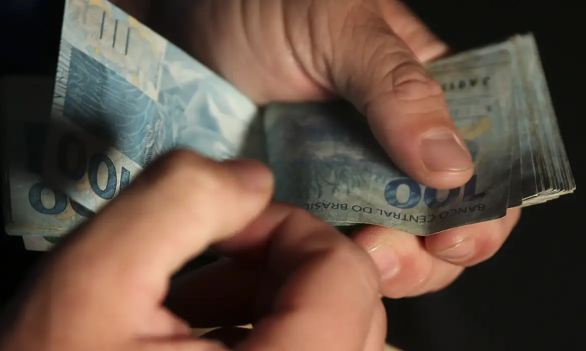 Mãos contando notas de cem reais simbolizando renegociação de dívidas pelo Desenrola Brasil