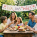 Família com três gerações sorri em volta de mesa rústica com bolos, frutas e suco, sob faixa escrita Dia das Mães.