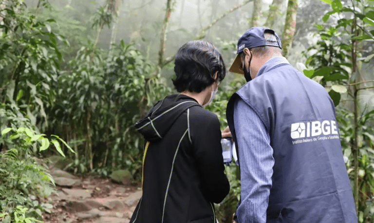 Dois servidores do IBGE com colete e boné institucionais em trabalho de campo dentro de área de mata