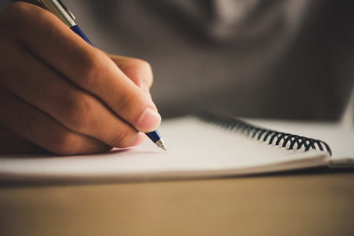 Mulher de camisa social escrevendo a lápis em caderno espiral em mesa de escritório com xícara e celular