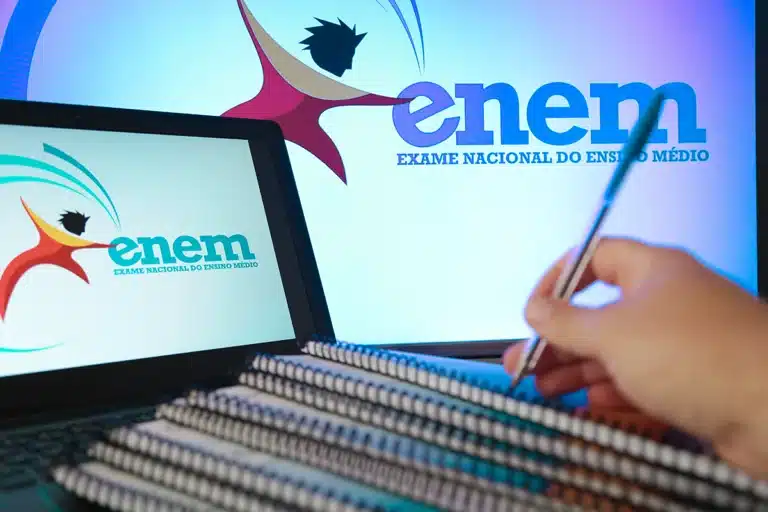 Mão segurando caneta sobre cadernos espirais, laptop exibindo logo do ENEM em ambiente de estudo azulado.