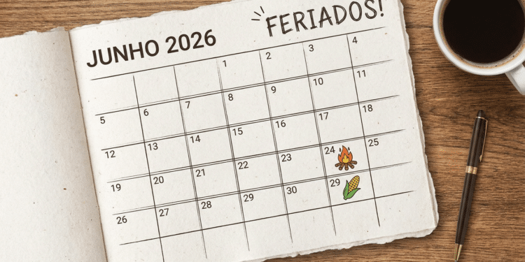 Calendário de junho 2026 sobre mesa de madeira com fogueira no dia 24 e milho no dia 29, xícara de café e caneta.