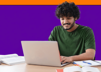 Estudante de cabelo cacheado sorri ao usar notebook sobre mesa com livros e cadernos abertos.