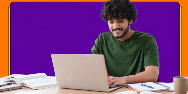 Estudante de cabelo cacheado sorri ao usar notebook sobre mesa com livros e cadernos abertos.
