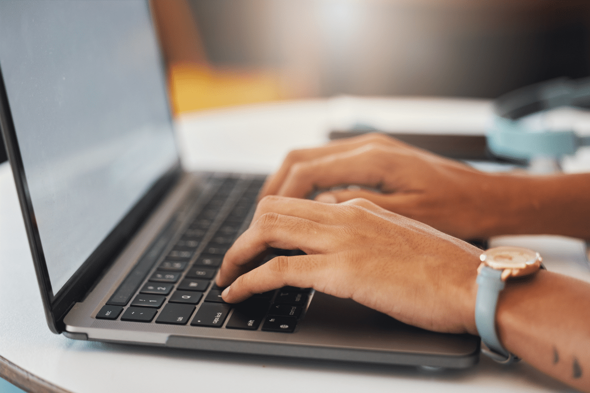 Mãos femininas digitam em teclado de notebook cinza, pulso com relógio dourado e pulseira azul turquesa.