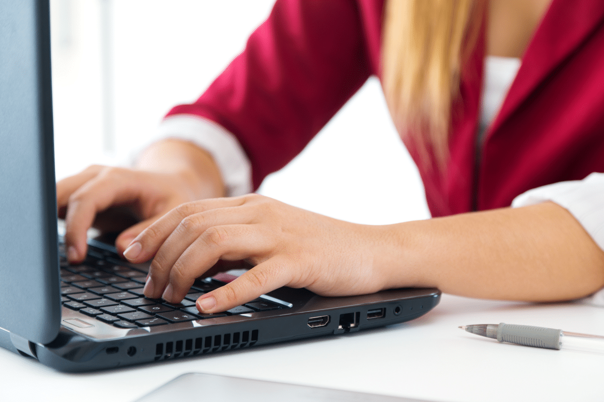 Mãos femininas digitando em teclado de notebook preto, blazer vermelho e cabelo loiro visíveis