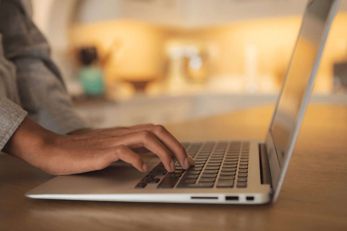 Mão feminina digitando em teclado de notebook prateado sobre mesa de madeira com iluminação quente ao fundo.