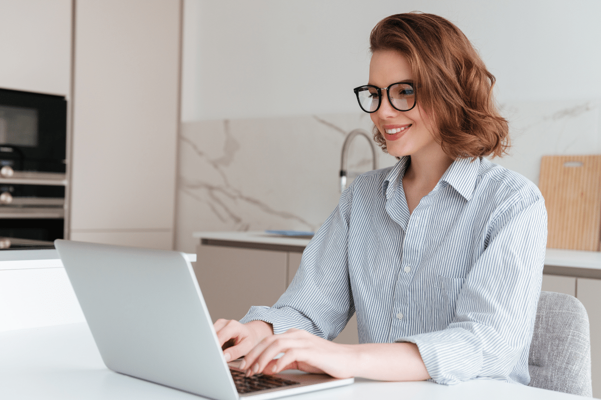 Mulher ruiva de óculos e camisa listrada digita sorrindo em laptop prateado na bancada de cozinha clara.