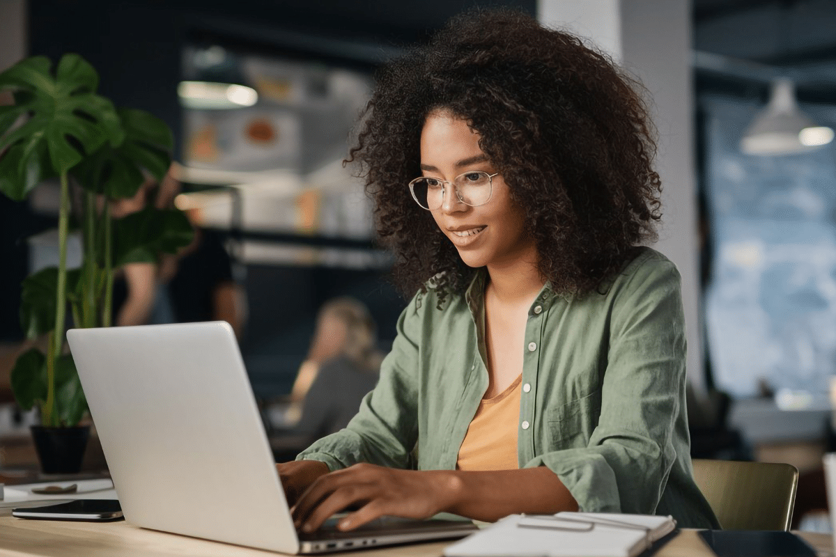 Mulher jovem de cabelos cacheados e óculos digita em laptop cinza em ambiente corporativo com plantas