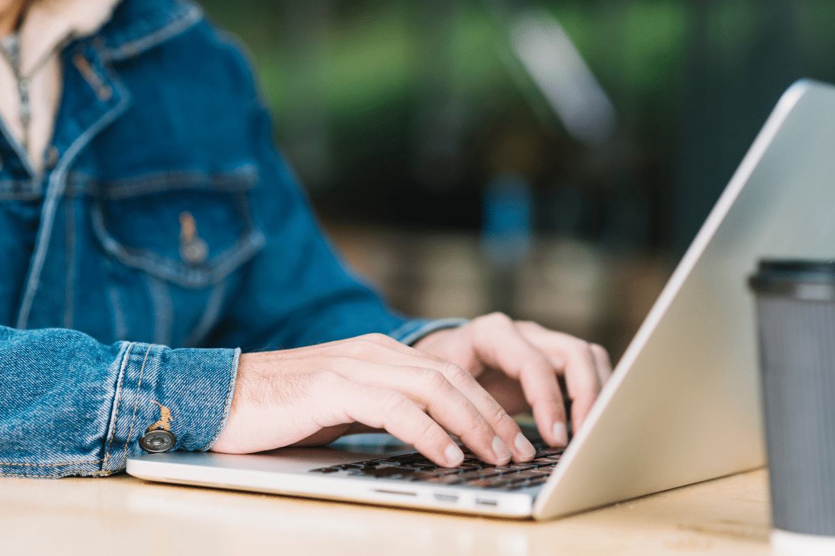 Mãos digitando em laptop prateado sobre mesa de madeira; pessoa usa jaqueta jeans azul, copo cinza ao lado.