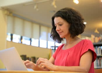 Mulher de blusa vermelha digita em notebook prateado sentada em ambiente de biblioteca com estantes de livros ao fundo