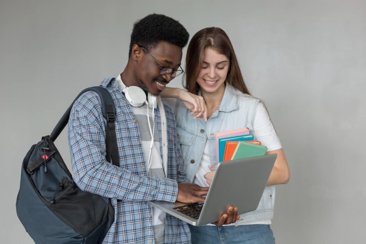Dois estudantes sorriem ao consultar um notebook enquanto a jovem segura cadernos coloridos em fundo neutro