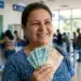 Mulher sorrindo segurando cédulas de real em agência da Caixa, representando a liberação de R$ 1.078,01.