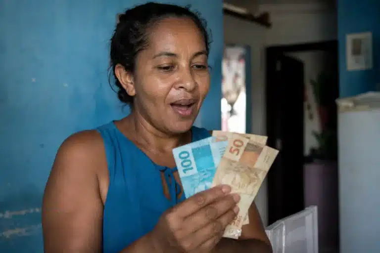 Mulher segurando cédulas de dinheiro e conferindo valor, representando o novo lote do auxílio de R$ 7,3 mil liberado pelo governo.