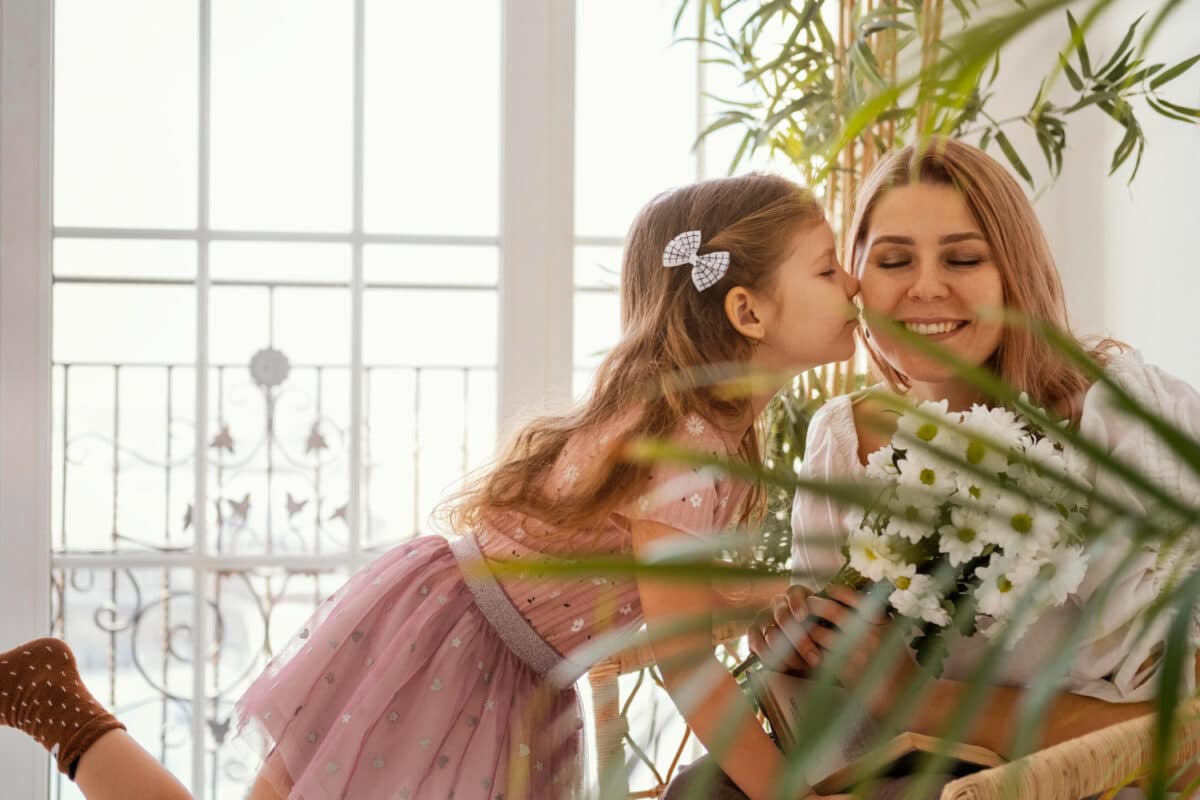 Menina beijando a mãe no rosto enquanto entrega um buquê de flores brancas