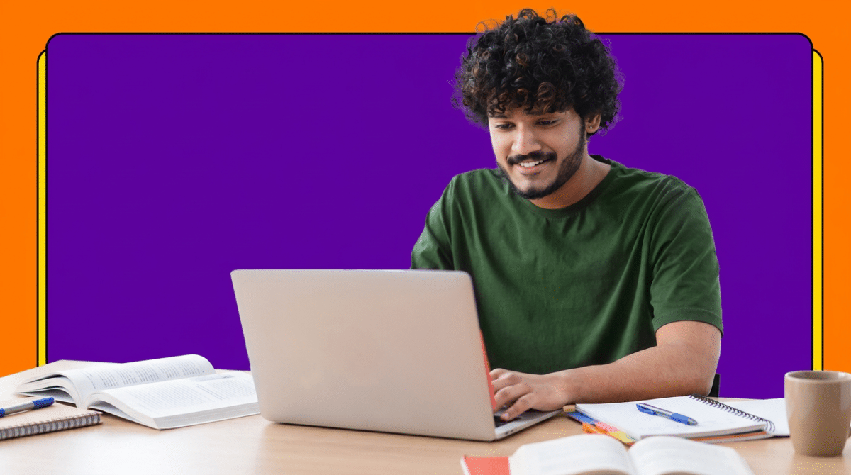 Jovem de camiseta verde sorri enquanto usa notebook sobre mesa com livros e cadernos de estudo.